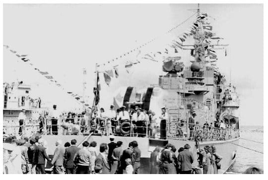 Ein über die Toppen geflaggtes Kriegsschiff liegt an der Pier, davor drängen sich Besucher an der Kaikante; an Deck stehen Besatzungsangehörige