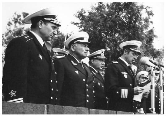 Mehrere Marineoffiziere in Ausgehuniform stehen auf einer Tribüne, einer von ihnen verliest am Mikrofon eine Ansprache