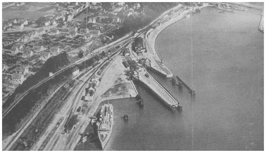 Luftbild des Saßnitzer Hafens mit Molen, Anlegebrücken, Gleisanlagen mit Güterwagen und der Stadtbebauung am Steilufer; an der Pier liegt ein Fährschiff