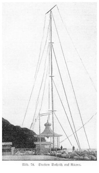 Historische Buchabbildung der Funkstation Saßnitz: ein hoher abgespannter Antennenmast über einem kleinen Stationshäuschen mit Zwiebelturm, davor zwei Männer