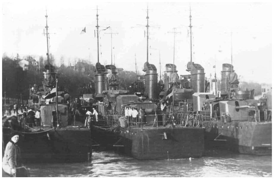 Vier Torpedoboote der Kaiserlichen Marine liegen im Päckchen im Saßnitzer Hafen, Besatzungsangehörige stehen an Deck, im Hintergrund bewaldetes Ufer mit Häusern