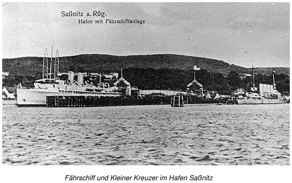 Historische Postkarte „Saßnitz a. Rüg. – Hafen mit Fährschiffanlage“: ein Fährschiff an der Anlegebrücke, dahinter am Kai ein Kleiner Kreuzer, im Hintergrund bewaldete Höhen