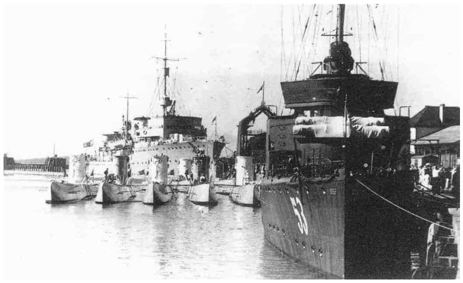 U-Boote liegen im Päckchen an der Pier des Saßnitzer Hafens, dahinter ein Begleitschiff; im Vordergrund ragt der dunkle Bug eines größeren Schiffes mit der Nummer 55 ins Bild