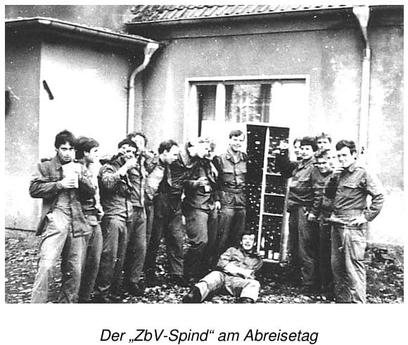 Eine Gruppe von Soldaten in Arbeitsuniform posiert lachend mit Trinkbechern vor einem Haus um ein Regal voller Flaschen, einer liegt am Boden